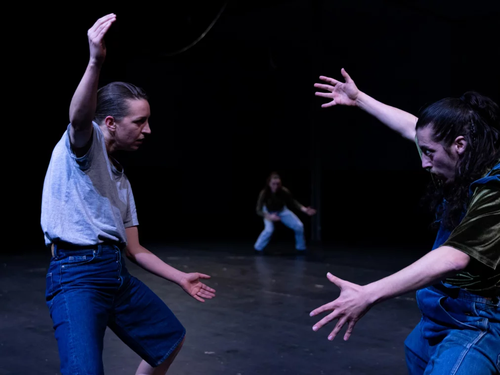 Drei Personen interagieren dynamisch auf einer dunklen Bühne. Zwei Personen stehen sich mit weit geöffneten Armen gegenüber, während eine weitere Person im Hintergrund in Bewegung ist. Kühles Bühnenlicht hebt die Gesten hervor.