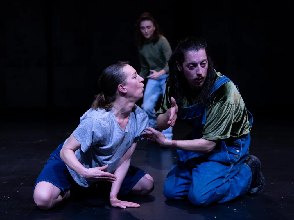 Zwei Personen knien sich gegenüber auf dem Bühnenboden. Eine Person beugt sich mit gespannter Körpersprache nach vorn, während die andere mit erhobener Hand reagiert. Eine dritte Person steht unscharf im Hintergrund.+IsabellaFernandesSantana©MayraWallraff-49