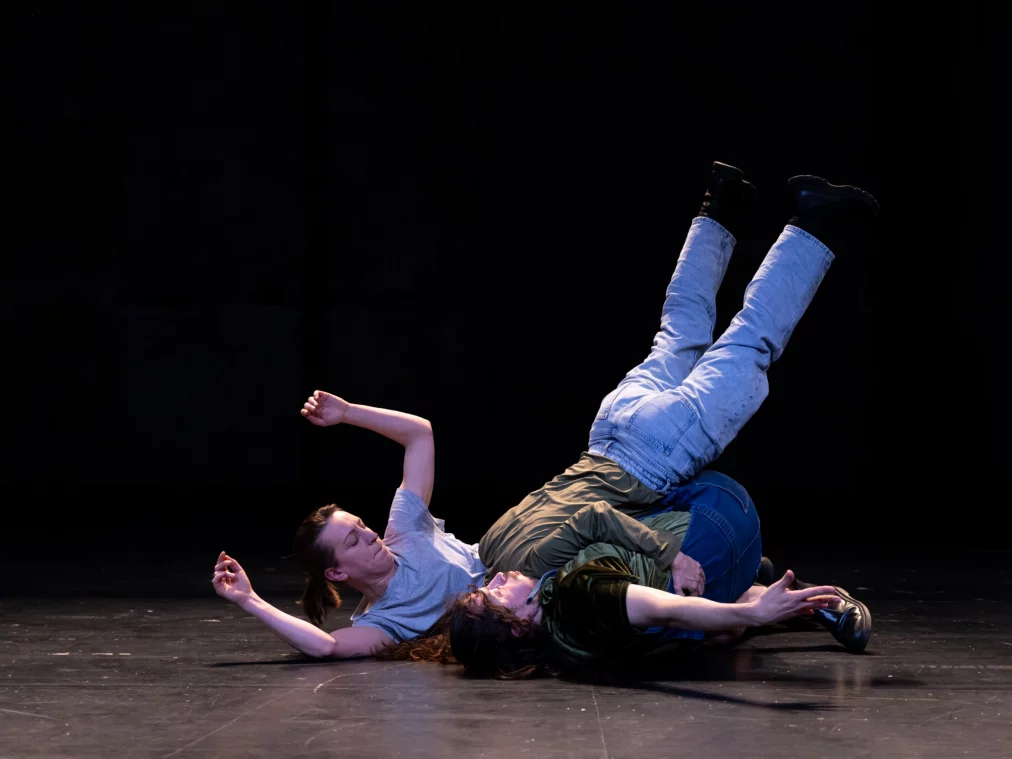 Zwei Personen liegen ineinander verschränkt auf dem Boden. Eine Person hat die Beine angehoben, während die andere flach auf dem Rücken liegt. Die Szene wirkt körperlich dicht und kraftvoll.
