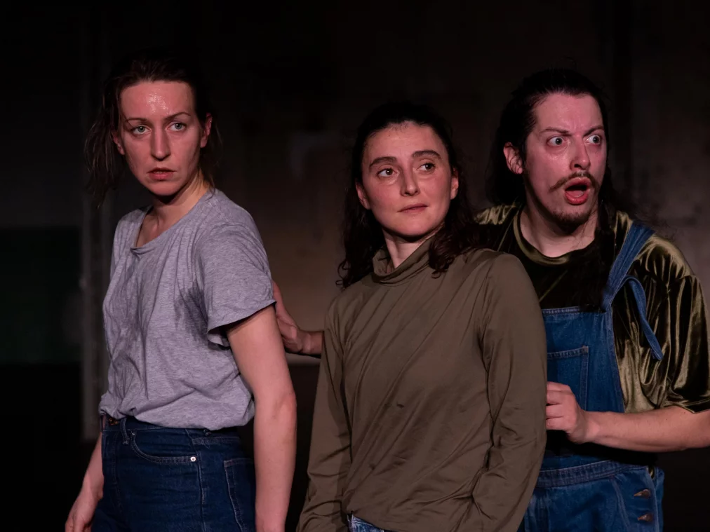 Drei Personen stehen dicht nebeneinander auf einer dunklen Bühne. Eine Person im grauen T-Shirt blickt angespannt zur Seite, eine Person in olivgrünem Oberteil schaut ruhig nach vorn, eine Person in Latzhose reagiert mit weit geöffnetem Mund. Gedämpftes Bühnenlicht betont die Gesichter.