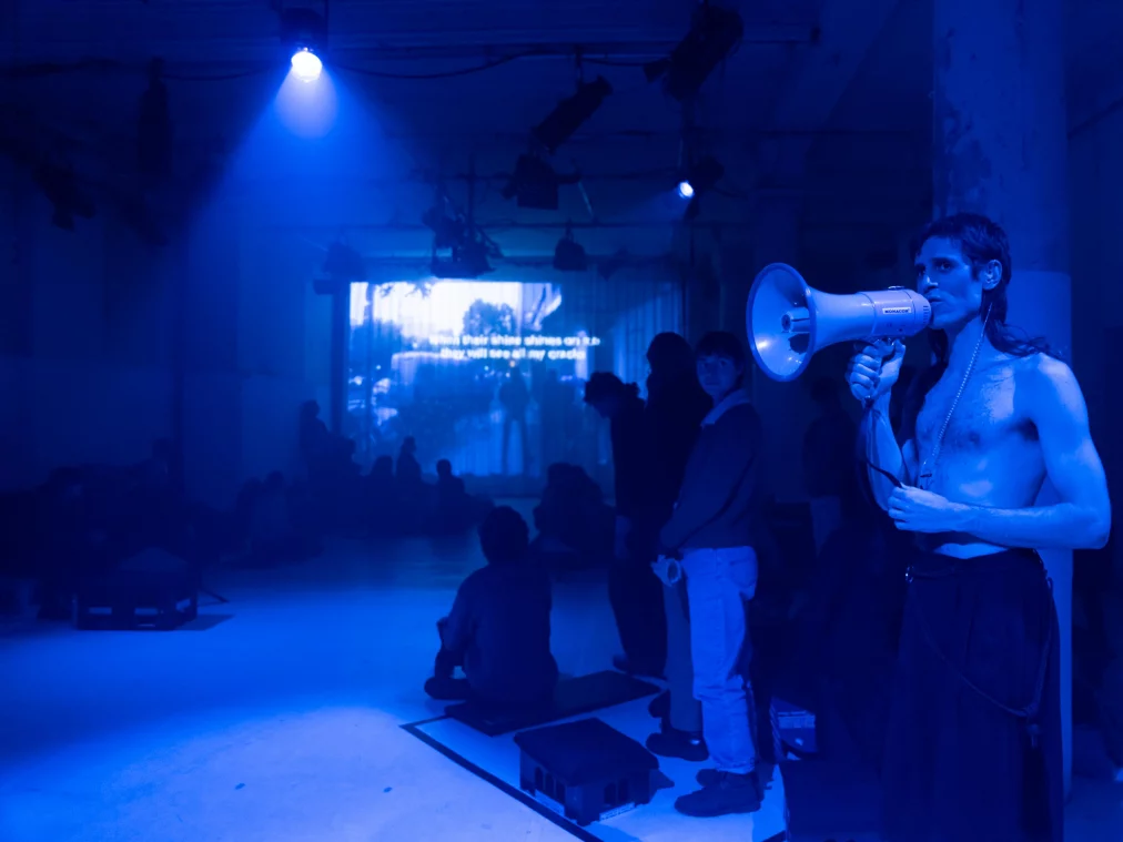 Eine obenrum nackte Person hält ein Megafon und steht seitlich im Raum unter blauem Licht. Im Hintergrund sind sitzende und stehende Personen sowie eine projizierte Textfläche zu sehen. Die Atmosphäre wirkt kühl und technisch.
