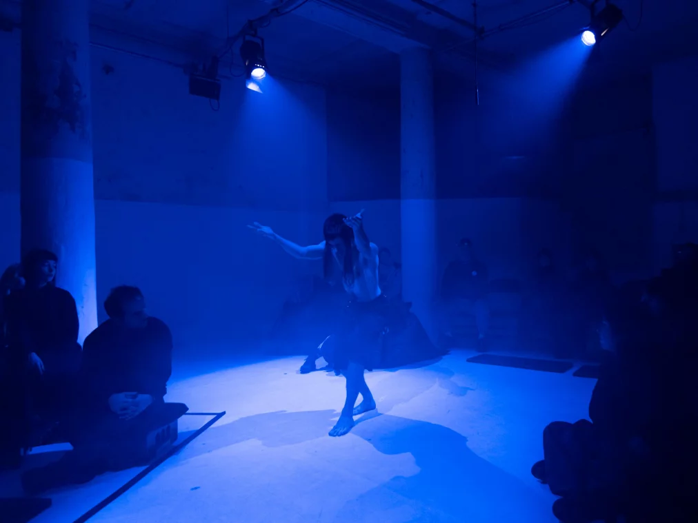 Eine barfuß stehende Person befindet sich im Zentrum eines Raumes, der in blaues Licht getaucht ist. Die Arme sind erhoben und leicht geöffnet. Rundherum sitzen Zuschauer*innen auf niedrigen Podesten und beobachten die Szene.