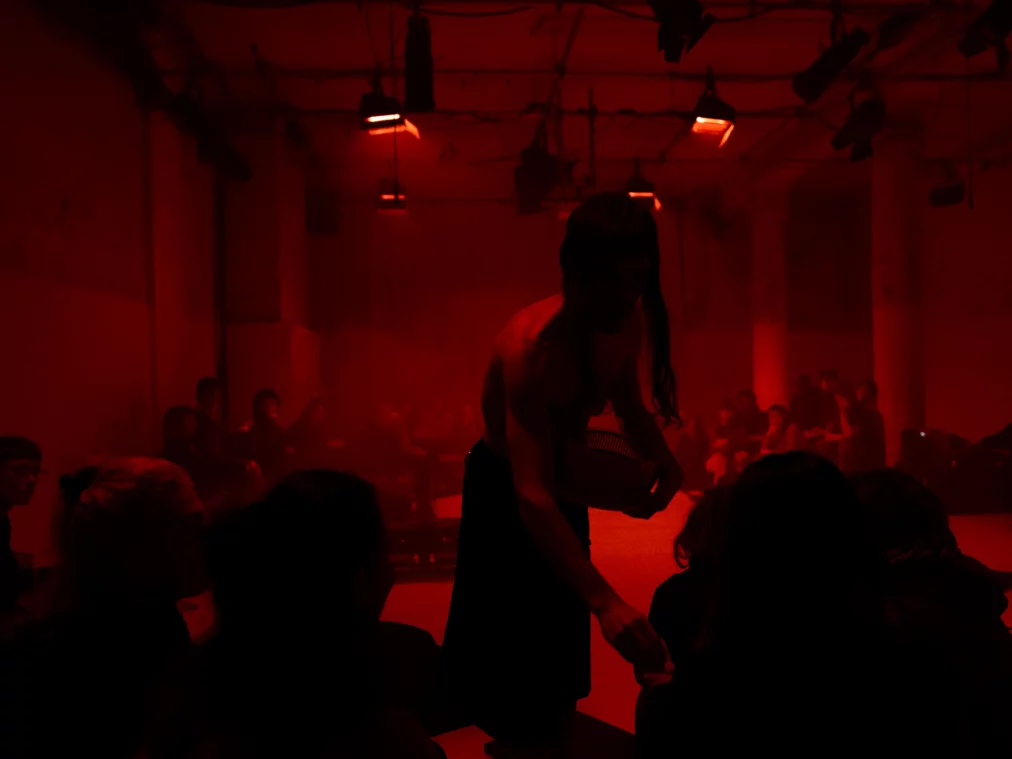 Eine obenrum nackte Person bewegt sich durch einen rot beleuchteten Raum mit Publikum. Der Oberkörper ist nach vorne geneigt, ein Objekt wird nah am Körper gehalten. Die Zuschauenden sitzen dicht um die Spielfläche herum.