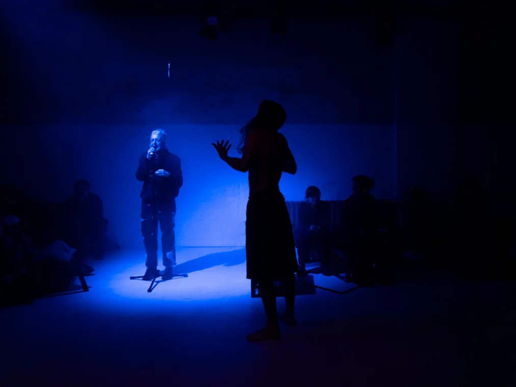 In blauem Licht steht eine Person am Mikrofon; davor zeichnet sich eine zweite Person als dunkle Silhouette mit erhobener Hand ab, während weitere Personen am Rand des Raums sitzen.