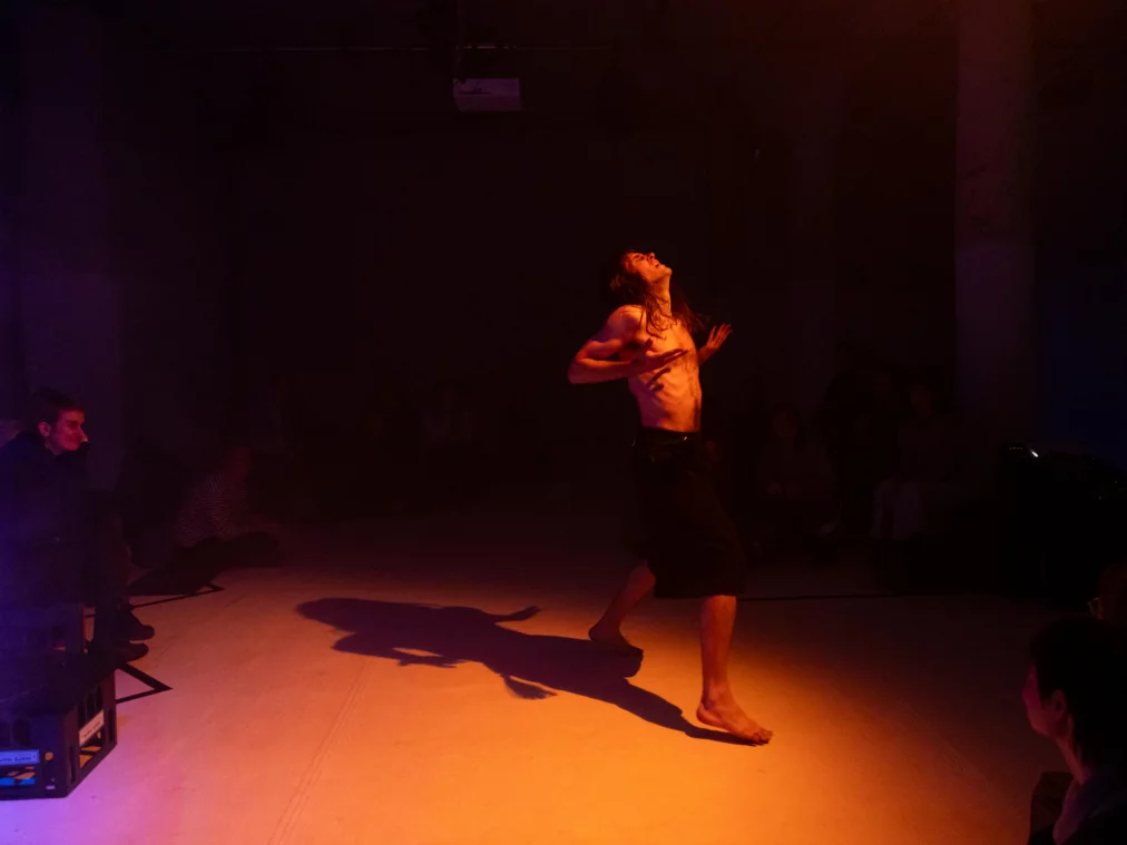 In warmem Rot-Orangeton bewegt sich eine oberkörperfreie Person barfuß über den Bühnenboden, den Kopf nach hinten geneigt und die Hände an die Brust geführt; ringsum sitzen Personen im Halbdunkel.