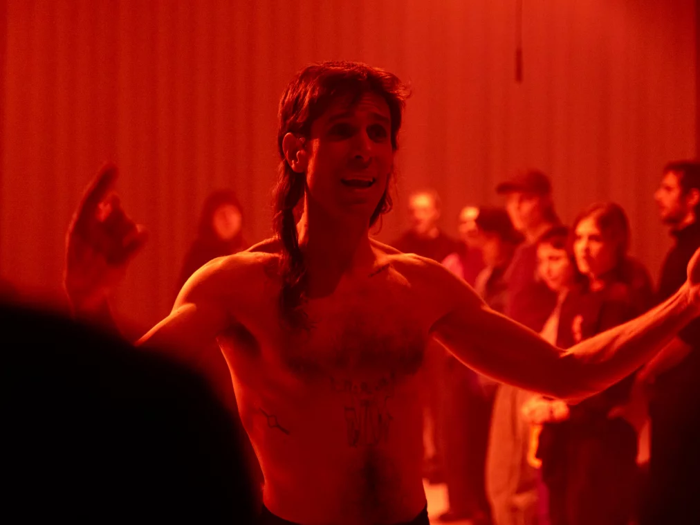 Eine obenrum nackte Person steht in rotem Licht zwischen sitzenden Zuschauer*innen. Die Arme sind weit ausgebreitet, der Mund ist geöffnet, als würde gesprochen oder gerufen. Die Umgebung wirkt dicht und theatral, mit roten Lichtquellen über der Szene.
