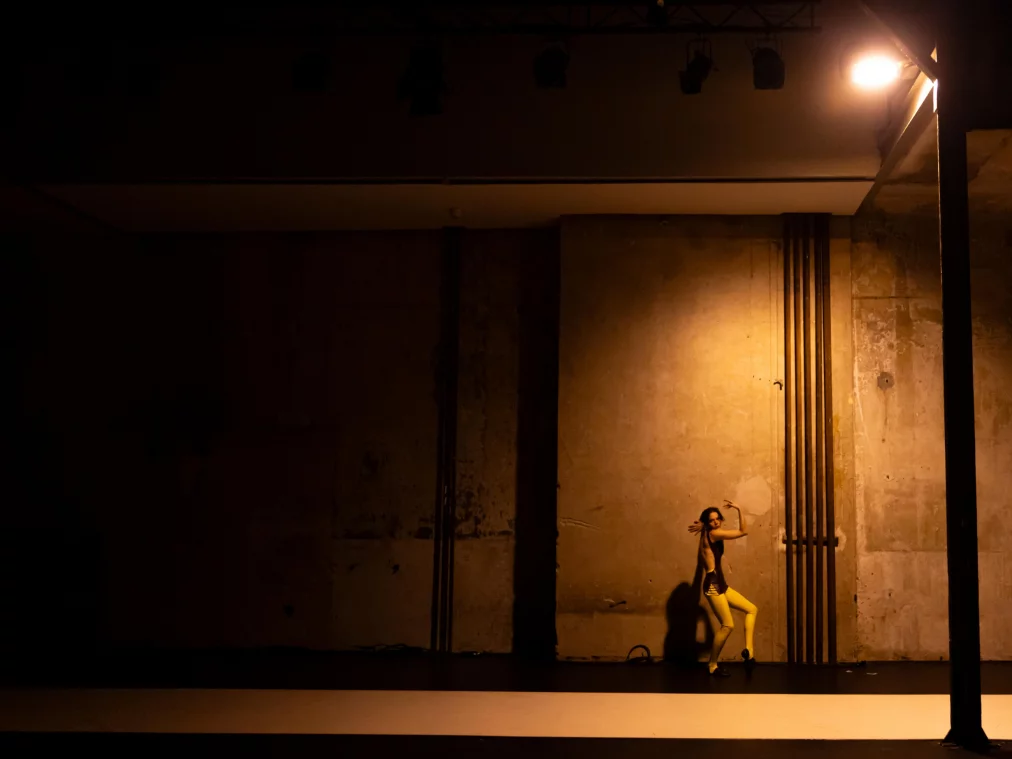 Eine einzelne Person steht weit entfernt vor einer hohen Wand in warmem Licht. Der Körper ist leicht gedreht, ein Arm hebt sich in einer präzisen, grafischen Bewegung.