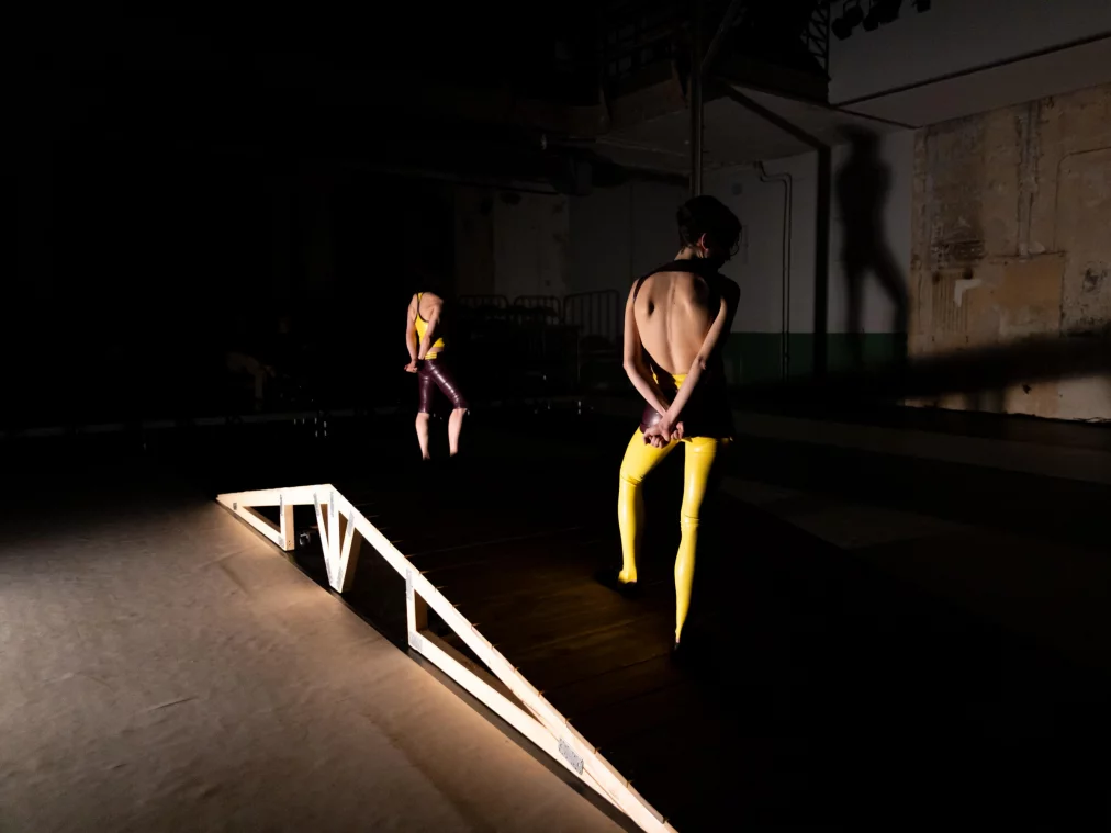 Zwei Personen befinden sich auf unterschiedlichen Ebenen einer Bühne mit Holzsteg. Eine Person steht im Vordergrund mit verschränkten Armen hinter dem Rücken, eine zweite Person bewegt sich weiter hinten im Halbdunkel.