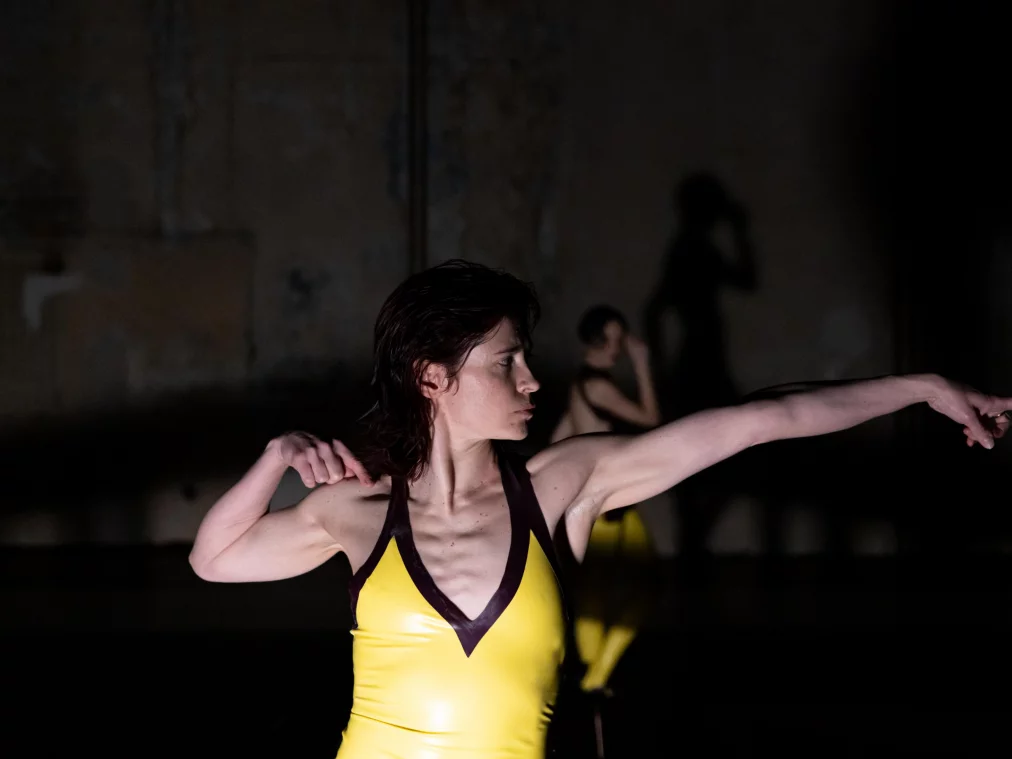 Eine Person im Vordergrund streckt einen Arm kraftvoll nach vorne und spannt den Oberkörper. Im Hintergrund ist eine zweite Person unscharf sichtbar, deren Schatten an der Wand erscheint.