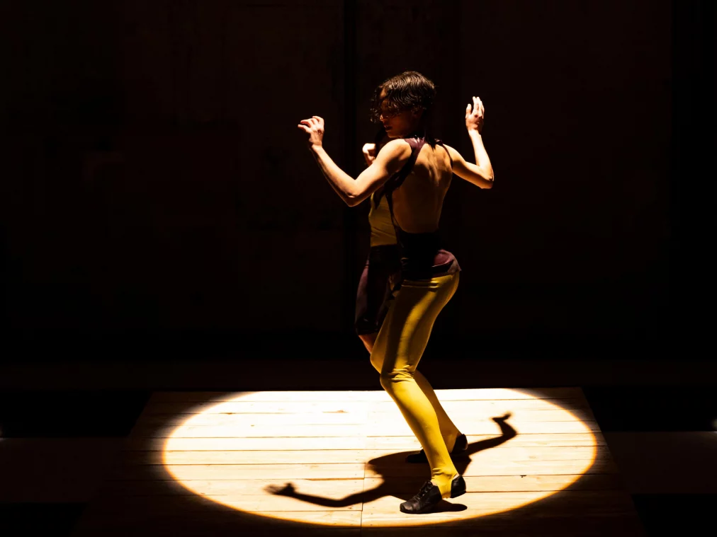 Eine einzelne Person steht mit dem Rücken zur Kamera auf einer erhöhten Holzfläche im runden Lichtkegel. Die Arme sind angewinkelt und angehoben, der Körper ist angespannt und leicht gedreht, während der Bühnenraum um die Figur dunkel bleibt.