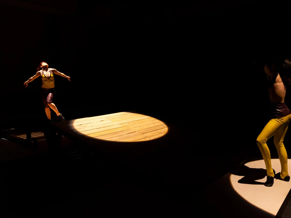 Zwei Personen stehen auf einer dunklen Bühne in getrennten Lichtkegeln. Eine Person bewegt sich mit ausgebreiteten Armen auf einer erhöhten Holzfläche, eine zweite Person steht seitlich im Licht und formt den Oberkörper in eine spannungsvolle Drehung.